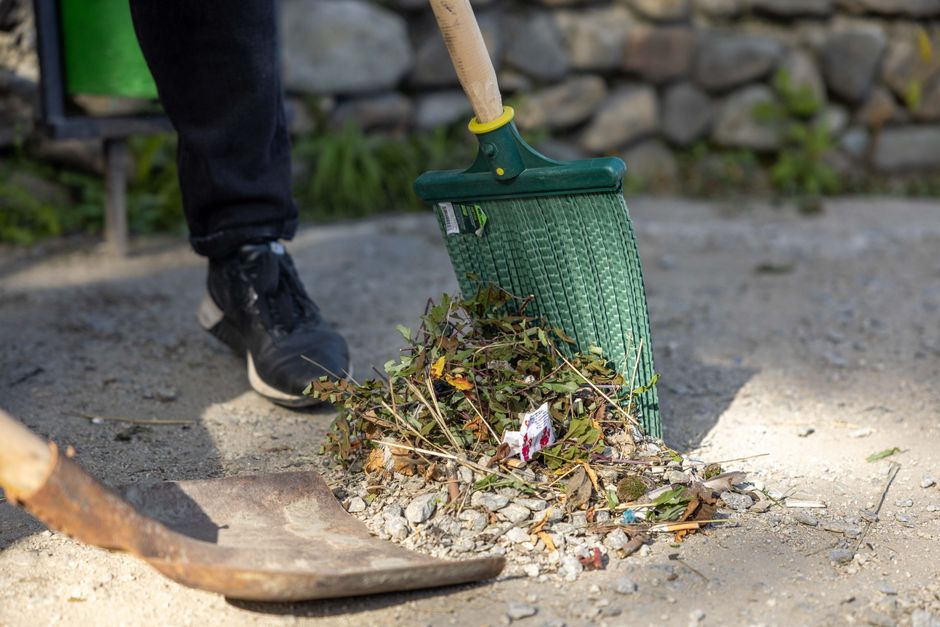 A person wearing black shoes and dark pants is using a green rake to clear fallen leaves, small twigs, and debris from a gravel pathway in a garden. The gravel surface is light gray with scattered lea